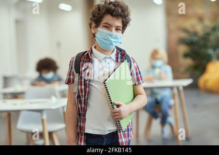 Portrait d'un jeune garçon d'école gai portant un masque pour empêcher la propagation de Covid19 regardant l'appareil photo, tenant un manuel en posant dans une salle de classe Banque D'Images