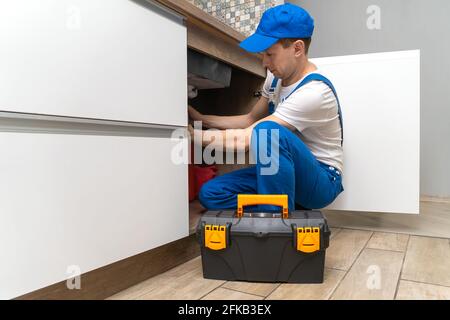 Le handyman en combinaison répare les tuyaux sous l'évier dans la cuisine, à côté d'une boîte d'outils Banque D'Images