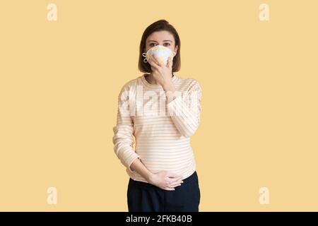 La mère enceinte caucasienne porte un masque médical posé sur un mur jaune de studio Banque D'Images
