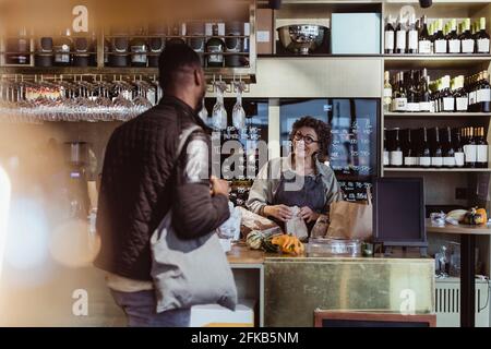 Une femme propriétaire de magasin parle avec un client masculin lors de l'emballage de la commande à la épicerie fine Banque D'Images