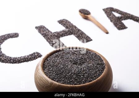 Mot chia orthographié avec des graines noires et une cuillère en bois, isolé sur la surface blanche avec un bol. Concept de manger propre. Supplément alimentaire végétalien, suralimentation. C Banque D'Images