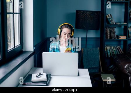 Une fille dans un casque sans fil est assise à une table avec un ordinateur portable dans le bureau à la maison. La fille tape sur le téléphone. Une jeune femme travaille intenamment de chez elle. Banque D'Images