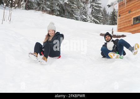 Jeune couple portant des vêtements d'hiver s'amusant et appréciant la promenade en traîneau dans un paysage instable. Paysage couvert de neige pendant la saison d'hiver avec des arbres. Une cabane en bois en arrière-plan. Banque D'Images