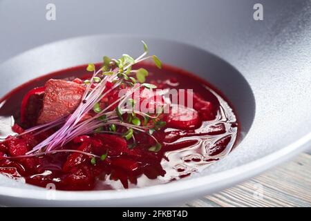 Vue rapprochée de borsch rouge vif, soupe de betteraves à la viande et aux légumes servis avec de jeunes pousses vertes. Délicieux plat chaud dans une assiette blanche au restaurant. C Banque D'Images