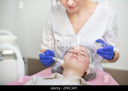 Cosmétologie et traitement de l'acné. Femme professionnelle dermatologue en gants de caoutchouc, applique le masque sur le visage du client avec la brosse de cosmétologie. Jeune fille de l'adolescence avec la peau problématique couché sur le canapé Banque D'Images