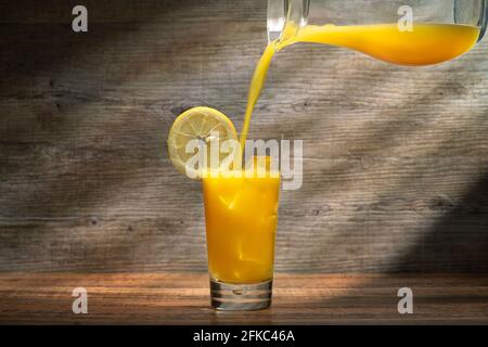 Jus d'orange frais versé dans un verre avec de la glace sur une table en bois avec des puits de lumière du soleil en arrière-plan. Banque D'Images