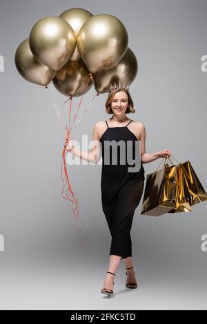 pleine longueur de femme heureuse de taille plus en noir slip robe et couronne tenant des ballons dorés et des sacs à provisions gris Banque D'Images