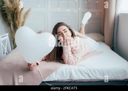 Une jeune fille rêveuse se trouve sur le lit et tient un ballon blanc en forme de coeur. L'amour et le concept de Saint Valentin. Banque D'Images