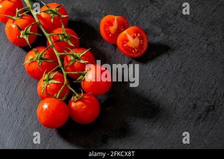 Belles tomates de vigne fraîches (cerise) avec des gouttes d'eau, brutes, entières et coupées en deux. Sur fond d'ardoise texturé noir. Ingrédients de la Méditerranée Banque D'Images