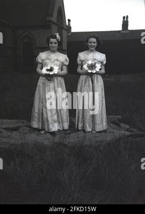 1958, historique, à l'extérieur, dans les jardins d'une église, deux adolescentes en robes longues et tenant des bouquets de fleurs, debout sur un tapis pour une photo avant le Carnaval de la Reine de mai. Ils accompagnent la Reine de mai, dans la procession, soit en marchant ou en faisant du vélo pendant les célébrations du jour de mai, une tradition ancienne qui a célébré l'arrivée du printemps et de l'été. Dans de nombreuses villes anglaises du nord, le défilé était relié aux écoles du dimanche de l'Église d'Angleterre. La figure de la reine de mai et sa pureté sont liées au culte et au folklore anciens. Banque D'Images