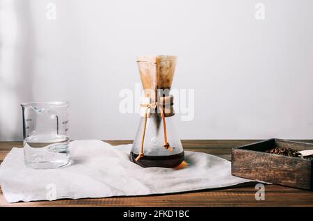 Cafetière manuelle en verre avec filtre et café sur un tissu gris sur une table en bois avec bac à grains de café et verre gradué sur les côtés Banque D'Images