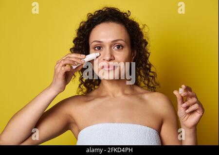 Portrait en gros plan d'une belle femme de beauté naturelle enveloppée une serviette de bain blanche contenant un tube de produit cosmétique et l'appliquer sous l'œil Banque D'Images