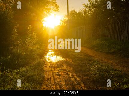 Coucher de soleil sur une route rurale dans le village après la pluie. Reflet du soleil dans les flaques. Banque D'Images