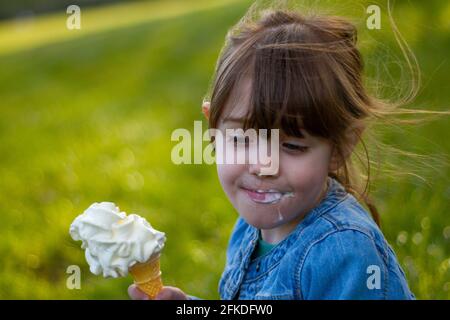 Gros plan d'une jolie fille aux cheveux bruns et aux yeux bleus, vêtue d'une veste en Jean bleu et mangeant une glace sur l'herbe par temps ensoleillé Banque D'Images