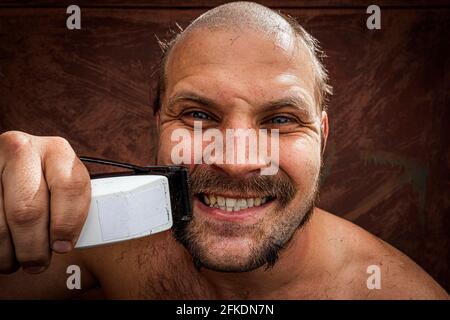 Homme joyeux adulte avec une barbe rasse sa barbe à la maison. Le gars regarde dans le miroir de salle de bains et utilise la tondeuse électrique. Vue avant Banque D'Images