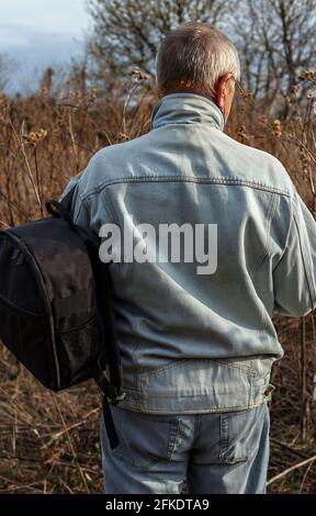 Homme âgé en costume denim avec sac à dos noir cherche à traverser les buissons tout en traversant les champs abandonnés. Banque D'Images