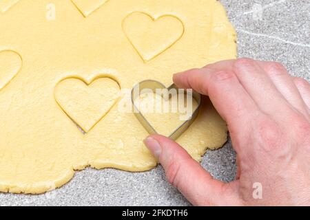 La main du chef pâtissier coupe des biscuits en forme de coeur de pâte jaune crue sur fond de table gris, gros plan. Concept de cuisson à domicile Banque D'Images