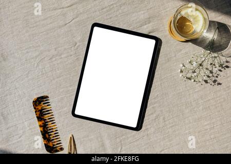 Maquette de tablette à écran vierge, verre d'eau avec citron, fleurs de gitsophila, peigne à cheveux sur la table avec nappe en lin. Flat lay, vue de dessus. Nordic, Scand Banque D'Images