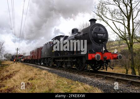 Train de fret à vapeur historique ou Loco puffant des nuages de fumée (conducteur de moteur sur la plaque de plancher, wagons de marchandises, homme côté piste) - KWVR, Yorkshire, Angleterre, Royaume-Uni. Banque D'Images