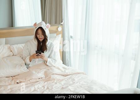 Jolie jeune femme en pyjama assise sur le lit après le réveil vérification des messages texte et des publications sur les réseaux sociaux Banque D'Images