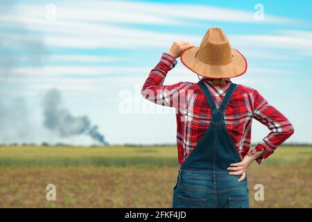Femme agricultrice concernée dans le champ de maïs regardant la fumée noire à l'horizon, concept d'assurance dans l'agriculture et l'agriculture Banque D'Images