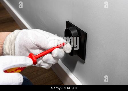 Un électricien utilise un tournevis rouge pour visser une prise électrique noire double mise à la terre dans le boîtier électrique de la salle. Banque D'Images