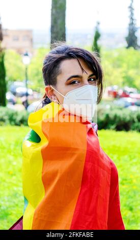Une femme transgenre / pas avec le drapeau gay sur ses épaules et un masque chirurgical. Personnes authentiques non binaires. Gay Pride Day pendant la pandémie de Covid Banque D'Images