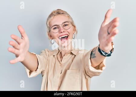 Belle femme caucasienne avec des cheveux blonds portant un look décontracté et des lunettes regardant la caméra souriant avec des bras ouverts pour la hug. Gai expression em Banque D'Images