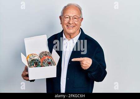 Homme âgé avec cheveux gris tenant pile de savoureux coloré boîte de beignets souriant, avec la main et le doigt Banque D'Images
