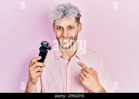 Jeune homme hispanique aux cheveux teints modernes tenant un rasoir électrique machine sourire heureux pointant avec la main et le doigt Banque D'Images