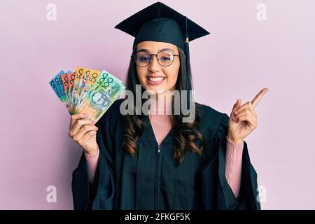 Jeune femme hispanique portant un uniforme de remise des diplômes tenant les dollars australiens souriants pointez avec votre main et votre doigt sur le côté Banque D'Images