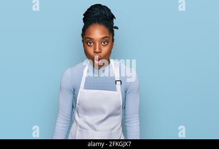 Jeune femme afro-américaine portant un tablier effrayée et stupéfaite de bouche ouverte pour le visage surprise, incrédulité Banque D'Images