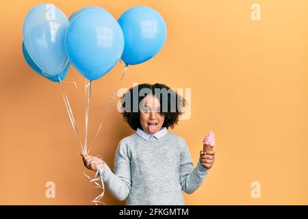 Jeune fille avec cheveux afro tenant la crème glacée et les ballons bleus collant la langue dehors heureux avec l'expression drôle. Banque D'Images