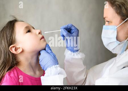 Test PCR COVID-19 et enfant, l'infirmière tient un écouvillon pour l'échantillon nasal d'un enfant adorable. Femme médecin en costume d'EPI examine la petite fille mignonne pendant le coronaviru Banque D'Images