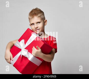 Portrait d'un garçon gai dans un t-shirt rouge tenant une grande boîte cadeau avec ruban dans les mains, le portant Banque D'Images