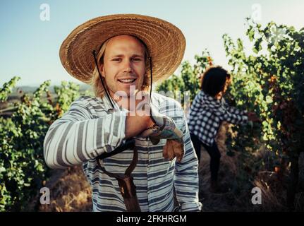 Mâle blond caucasien fermier souriant dans les vignobles portant chapeau de paille s'appuyer sur la fourche de pas Banque D'Images