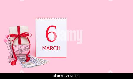 6e jour de mars. Une boîte cadeau dans un chariot, des dollars et un calendrier avec la date du 6 mars sur fond rose. Mois de printemps, jour du yea Banque D'Images