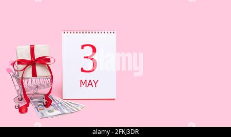 3e jour de mai. Une boîte cadeau dans un chariot, des dollars et un calendrier avec la date de 3 mai sur fond rose. Printemps mois, jour de l'année co Banque D'Images