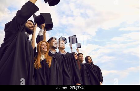 Les diplômés vêtus de manteau noir debout contre un ciel lumineux avec des chapeaux de remise des diplômes surélevés. Banque D'Images