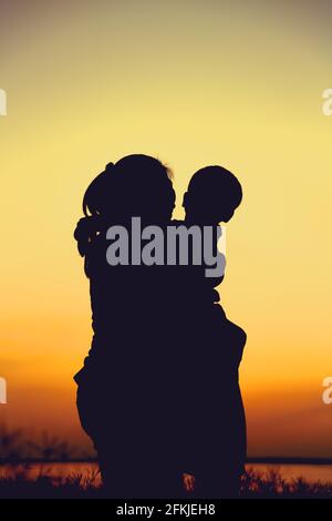 Silhouette de mère et d'enfant en profitant de la vue au bord de la rivière. Mère embrassant son fils sur fond de ciel de coucher de soleil coloré. Famille sympathique. Banque D'Images