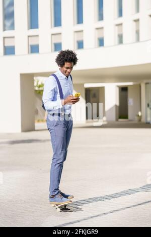 Homme d'affaires noir sur un skateboard regardant son smartphone à l'extérieur. Banque D'Images