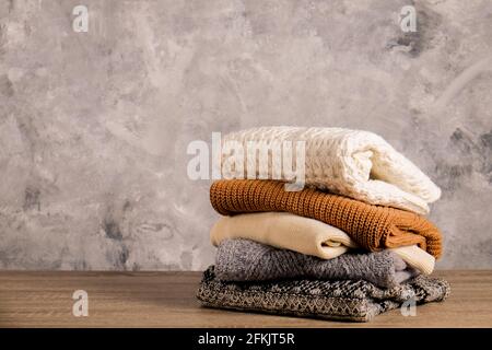 Ensemble de chandails en tricot doux couleur pastel, différents motifs de tricot pliés dans une pile désordonnée sur une table en bois brun, fond de mur de texture de béton Banque D'Images