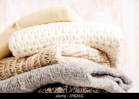 Lot de chandails en tricot doux couleur pastel, différents motifs de tricot pliés dans une pile désordonnée sur une table en bois brun, fond de mur à texture grunée. Banque D'Images