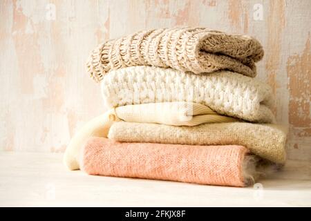 Lot de chandails en tricot doux couleur pastel, différents motifs de tricot pliés dans une pile désordonnée sur une table en bois brun, fond de mur à texture grunée. Banque D'Images