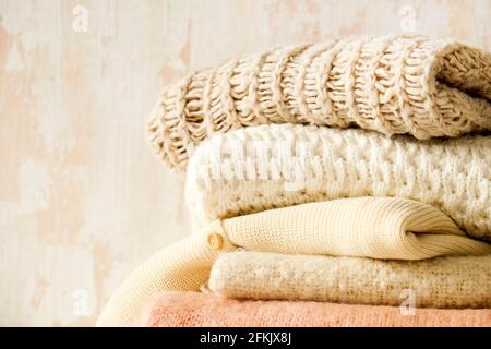 Lot de chandails en tricot doux couleur pastel, différents motifs de tricot pliés dans une pile désordonnée sur une table en bois brun, fond de mur à texture grunée. Banque D'Images