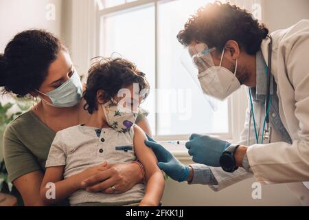 Une travailleuse de première ligne qui injecte le vaccin Covid-19 à un petit garçon assis avec sa mère à la maison. Enfant se faisant vacciner à la maison pendant une pandémie. Banque D'Images