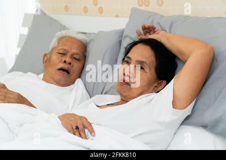Femme asiatique plus âgée, allongé les yeux ouverts dans le lit à côté de son mari qui ronflent et fait du bruit, des problèmes maritaux. Banque D'Images