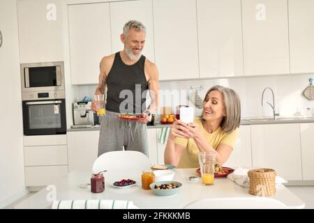 Un couple de familles âgées heureux prenant le petit déjeuner au téléphone dans la cuisine. Banque D'Images
