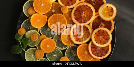 Citruses fraîches sur plaque ronde sur fond noir foncé en pierre, couché à plat. Oranges, clémentines, mandarines, limes. Vue de dessus. Cuisine spectaculaire Banque D'Images