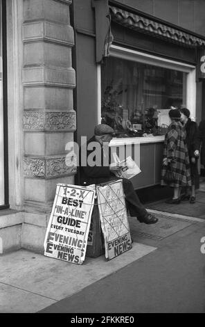 années 1950, hsitorical, un vieux vendeur de journaux mâles avec du papier en main et portant un chapeau de tissu assis à l'extérieur sur un trottoir. The Headline in the Evening News, « 15,000 Reds in Big Korea Battle », Londres, Angleterre, Royaume-Uni. La guerre de Corée a eu lieu entre juin 1950 et juillet 1953. Banque D'Images